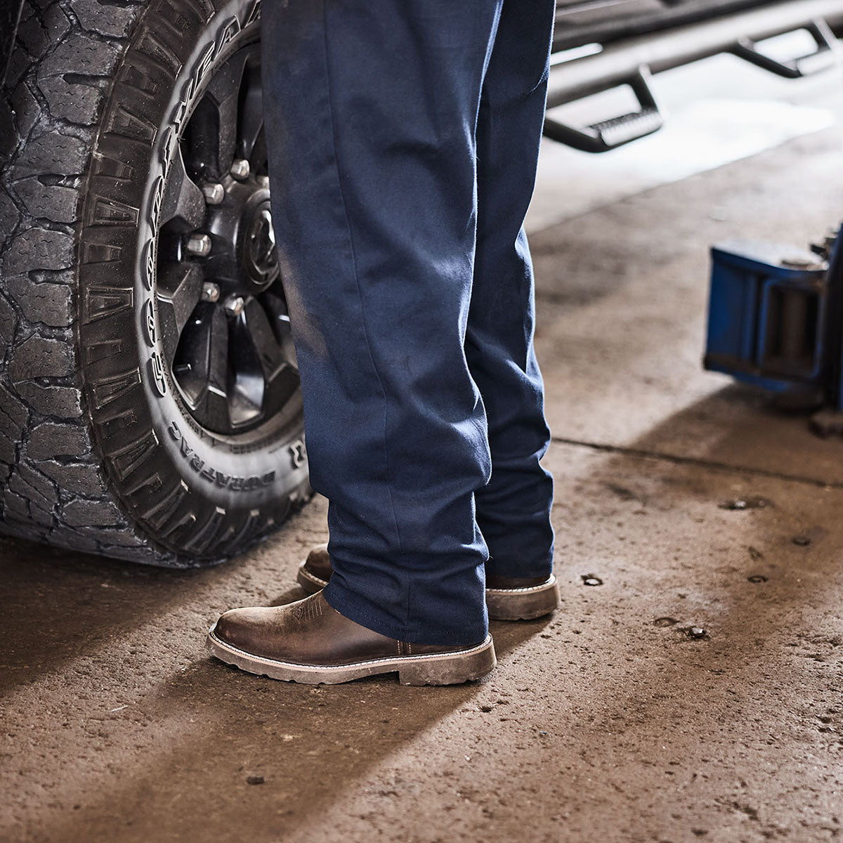 Person wearing blue pants and brown shoes standing next to a tire in a garage.